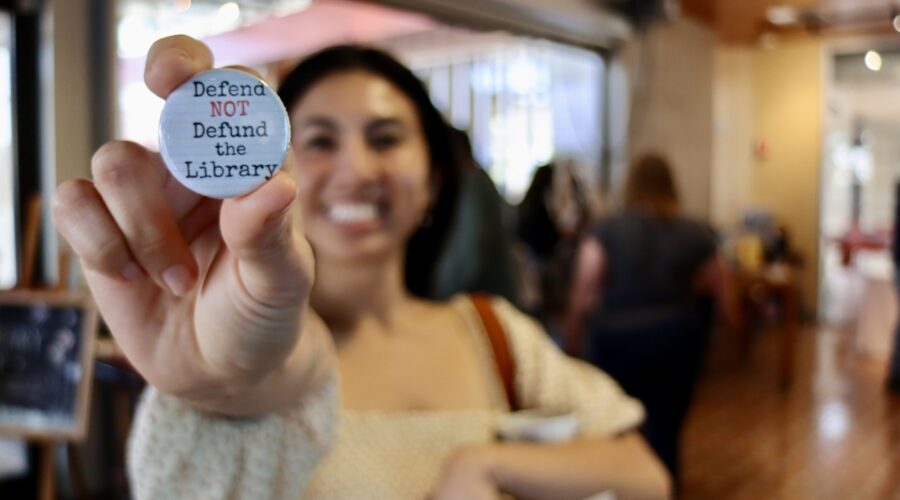 Image contains: A San Diego Book Crawler holds up a 'Defend NOT Defund the Library' pin.