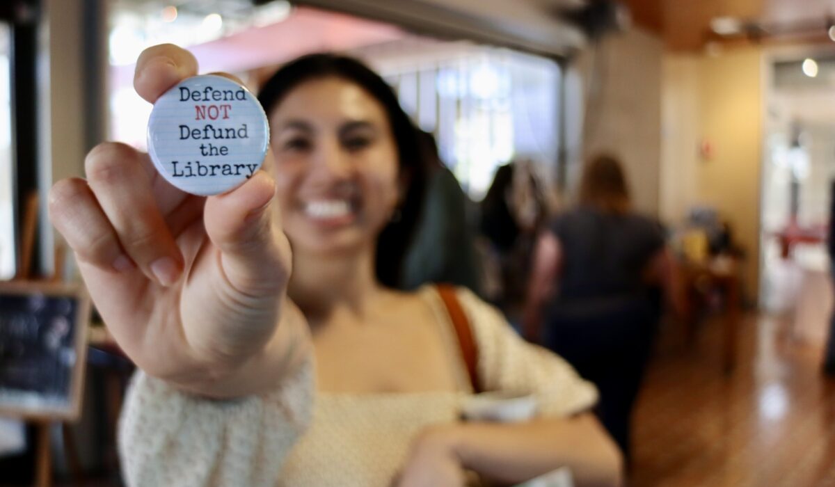 Image contains: A San Diego Book Crawler holds up a 'Defend NOT Defund the Library' pin.